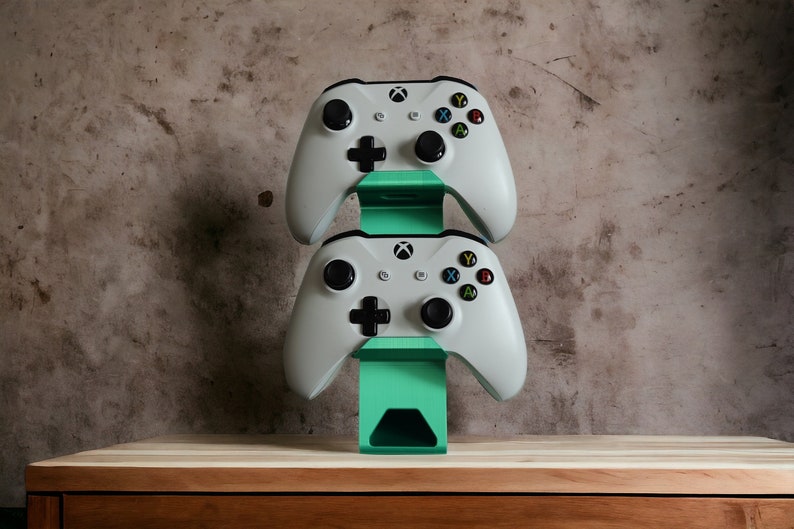 K&ouml;nnte beinhalten: Zwei wei&szlig;e Xbox-Controller werden auf einem gr&uuml;nen St&auml;nder angezeigt. Der St&auml;nder steht auf einer Holzoberfl&auml;che.