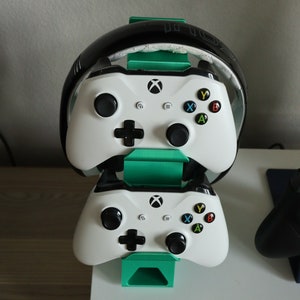 K&ouml;nnte beinhalten: Zwei wei&szlig;e Xbox-Controller sind auf einem gr&uuml;nen St&auml;nder gestapelt, mit einem schwarzen Headset oben drauf. Ein schwarzer Xbox-Controller befindet sich rechts auf einem blauen St&auml;nder.