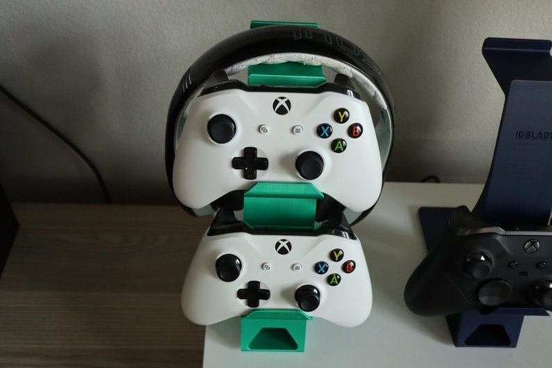 K&ouml;nnte beinhalten: Zwei wei&szlig;e Xbox-Controller sind auf einem gr&uuml;nen St&auml;nder gestapelt, mit einem schwarzen Headset oben drauf. Ein schwarzer Xbox-Controller befindet sich rechts auf einem blauen St&auml;nder.