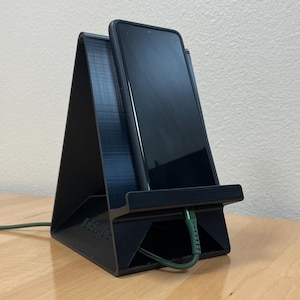 May include: A black triangular smartphone stand with a textured finish holds a dark smartphone in an upright position. A green charging cable is plugged into the bottom of the device, passing through the base of the stand.
