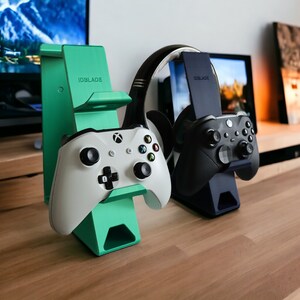 K&ouml;nnte beinhalten: Zwei Gaming-Controller-St&auml;nder, einer gr&uuml;n und einer schwarz, mit Controllern und einem Headset oben drauf. Die St&auml;nder sind mit "IDBLADE" beschriftet.