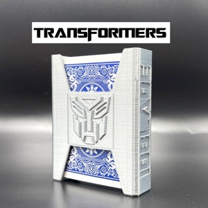 May include: A gray plastic card holder with a Transformers logo on the front. The card holder is designed to hold a standard deck of playing cards.