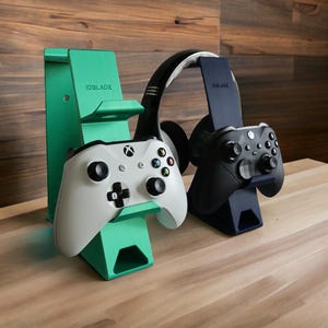May include: A mint green and a navy blue gaming stand, each featuring the text IDBLADE. The mint stand holds a white game controller, while the navy stand holds a black game controller and a pair of black and white over-ear headphones.