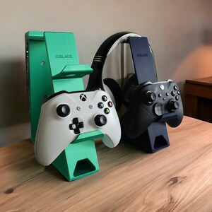 K&ouml;nnte beinhalten: Zwei gr&uuml;ne und schwarze Gaming-Controller-St&auml;nder mit einem wei&szlig;en und schwarzen Controller auf jedem St&auml;nder. Die St&auml;nder haben den Schriftzug "IDBLADE" aufgedruckt. Die St&auml;nder stehen auf einer Holzoberfl&auml;che.