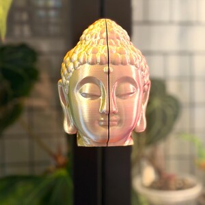 May include: A two-piece Buddha head sculpture with a pearlescent finish. The sculpture is split down the middle, revealing the face of Buddha with closed eyes. The head is a light pink and gold color, with a detailed textured design.