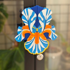 May include: A colorful, floral-shaped magnet in shades of blue, white, orange, and yellow. The design resembles an iris flower, with detailed petal patterns. The magnet is attached to a dark surface, likely a door or cabinet.