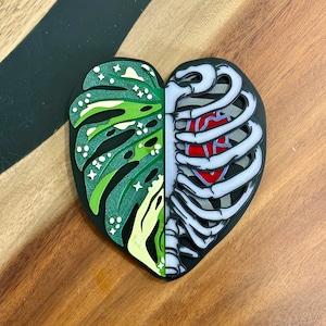 May include: A heart-shaped resin pin with a green and white monstera leaf on one side and a black and white ribcage with a red heart on the other side.