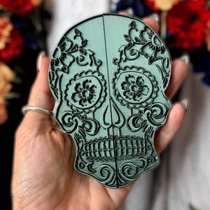 May include: A light mint green skull-shaped object featuring intricate black embossed floral and scrollwork patterns. The design includes detailed eye sockets with flower motifs and a defined jawline, split vertically into two symmetrical halves.