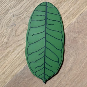 May include: A green leaf-shaped coaster with a detailed vein pattern. The coaster is made of a textured material and has a slightly rounded shape.