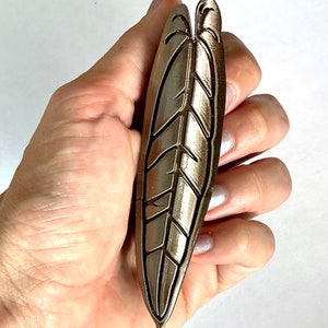 Könnte beinhalten: Ein metallischer goldfarbener Anhänger in Blattform mit einem detaillierten Design. Der Anhänger wird in einer Hand gehalten.