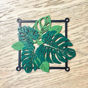 May include: A decorative wall art piece featuring green Monstera leaves within a black square frame. The leaves have intricate cut-out details and varying shades of green. The frame has small circular accents at each corner. The artwork is set against a light wood grain background.