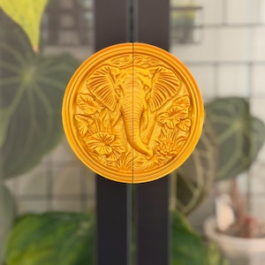 May include: A pair of golden door handles, each featuring a detailed relief of an elephant surrounded by floral elements. The handles are circular and mounted on a dark, vertical frame, creating a decorative and functional design.