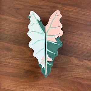 May include: A two-piece decorative object shaped like a variegated Alocasia leaf. The left piece features white and light green sections, while the right piece displays light pink and dark green glittery textures with defined leaf veins.