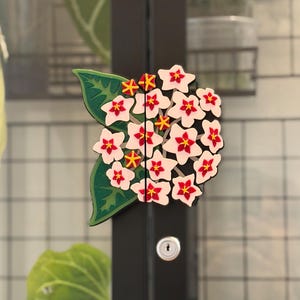 May include: A decorative cabinet handle featuring a cluster of white Hoya flowers with red centres and yellow star-shaped accents, accompanied by a large green leaf. The floral design is split into two halves, mounted on a black vertical frame.