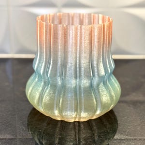 May include: A decorative vase with a gradient color scheme, transitioning from orange to blue and green. The vase has a ribbed design and a rounded base, suitable for home decor.