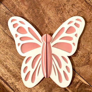 May include: A white butterfly wall decor with pink accents. The butterfly has intricate cutouts and a 3D design.