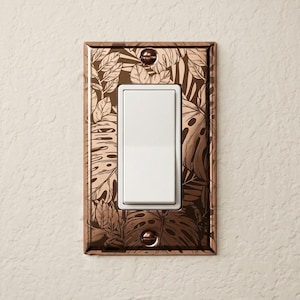 May include: A rectangular light switch cover plate featuring a metallic copper finish with a dark brown botanical leaf pattern. The plate has a central vertical white rocker switch and is secured by two matching copper screws at the top and bottom.