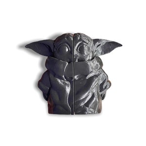May include: A black 3D printed figurine of a character from the Star Wars franchise, known as "The Child" or "Baby Yoda". The figurine is posed with its arms crossed and has a detailed face with large eyes.