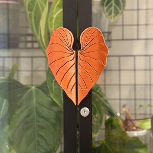 May include: A pair of orange, heart-shaped cabinet handles with a textured, leaf-like vein pattern. The handles are mounted vertically on a black metal frame, positioned directly above a small, circular silver keyhole lock mechanism.