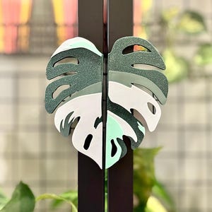 May include: A green and white monstera leaf cutout, painted with a speckled texture, is attached to a black metal rod.