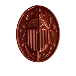 May include: A copper-colored, oval-shaped pendant with a detailed design of a scarab beetle. The beetle has a raised, textured body and intricate wings.