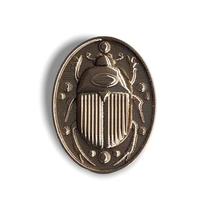 May include: A silver-toned metal pin with an oval shape. The pin features a detailed design of a scarab beetle with a raised, textured surface.