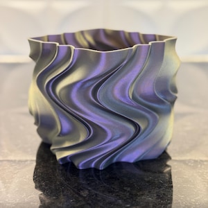 May include: A decorative, iridescent planter with a wavy, textured design. The planter has a square shape with a ruffled top edge. The colors shift from purple to silver and gold, creating a unique visual effect. The planter is sitting on a dark surface.