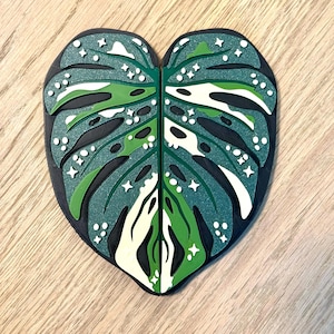 Könnte beinhalten: Ein grünes und weißes Monstera-Blatt mit glitzernder Oberfläche. Das Blatt ist herzförmig und hat einen schwarzen Umriss.