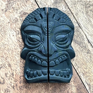 May include: A black tiki mask carved in a traditional style. The mask is split into two halves, each with a detailed face and a wide, toothy grin.