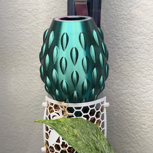 Op de afbeelding: Een tealgroene keramische plantenpot met een getextureerd, stekelig ontwerp. De pot hangt aan een witte metalen houder met een honingraatpatroon.