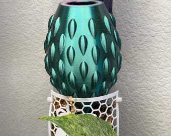 Op de afbeelding: Een metallic blauwgroene, eivormige houder met een gestructureerd, verhoogd druppelpatroon aan de buitenkant. Het object is geplaatst boven een witte, cilindrische plantensteun met honingraatpatroon die bruin mos en een groen blad bevat.