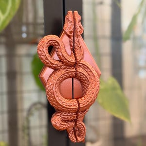 May include: A copper-colored door handle featuring a detailed, textured snake coiled into a circular shape. The snake is split vertically down the center, mounted onto a diamond-shaped backplate. The metallic finish has a subtle sheen.