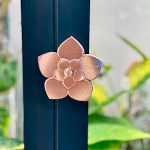 Può includere: Un fiore di metallo color rame con cinque petali è attaccato a un palo di recinzione in metallo blu scuro. Il fiore ha una superficie testurizzata e un centro leggermente rialzato.