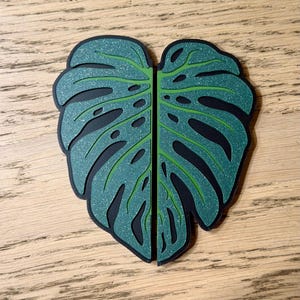 May include: A two-piece decorative object shaped like a split monstera leaf. The leaf features a dark green glittery finish on the top layer with cut-out sections revealing a solid black base layer underneath, creating a layered effect.