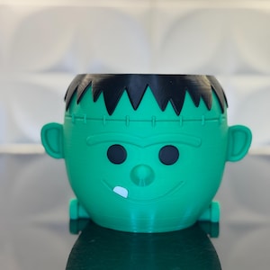 May include: Green Frankenstein-themed planter with black hair and a toothy grin.