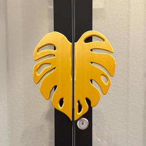 以下が含まれることがあります： 銀色の鍵穴が付いた黄色のモンステラ葉の形をしたドアノブ。ノブは黒いドアに取り付けられています。