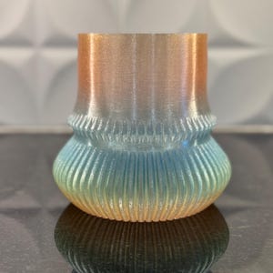 Può includere: Un piccolo vaso di vetro iridescente con un design testurizzato e nervato. Il vaso presenta un gradiente di colori, che parte da un arancio chiaro in alto e passa a un blu chiaro in basso.