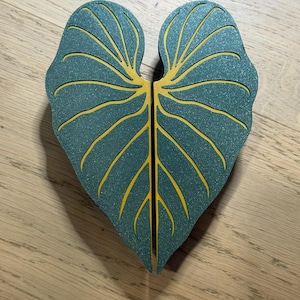 May include: A pair of heart-shaped decorative objects featuring a teal, glittery finish with recessed yellow veins. The two pieces are positioned side-by-side to form a single heart shape, showcasing a smooth texture and distinct leaf-like patterns.
