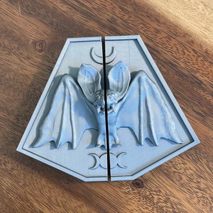May include: A gray, 3D printed, two-piece wall decor featuring a bat with outstretched wings and a crescent moon above it. The bat is centered between two triple moon symbols.