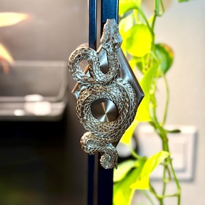 May include: A bronze-colored, decorative snake sculpture with intricate detailing. The snake is coiled around a diamond-shaped base, mounted on a black metal rod. The sculpture is set against a blurred background with green foliage.