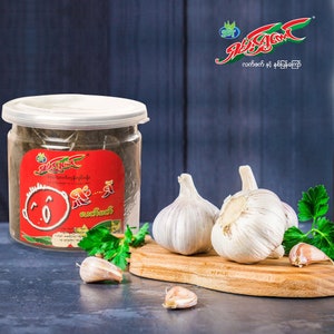 May include: A clear jar with a red label and a cartoon character on it. The label says "အရသာရှိတဲ့ ကြက်သွန်ဖြူ ဟင်းချက်အတွက်". The jar is sitting on a table with garlic cloves and parsley.