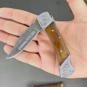 以下が含まれることがあります： 木製のハンドルと彫刻された金属のアクセントが付いた折りたたみ式のポケットナイフ。 刃は部分的に開いており、「Groomsman 28/03/2026」の文字が表示されています。 ナイフの下には、「Love You」という文字が入った茶色の革ケースが見えます。