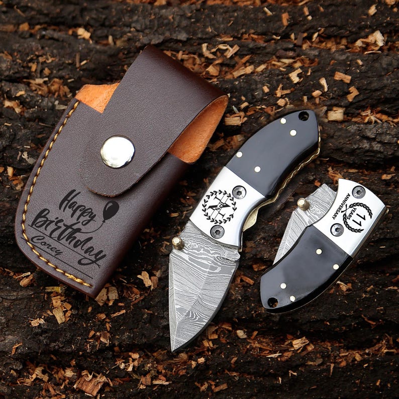 May include: A brown leather knife sheath with the words "Happy Birthday Corey" and a balloon graphic. Two folding knives with black and silver handles and patterned blades are next to the sheath. One knife has an "11 Years Anniversary" emblem.
