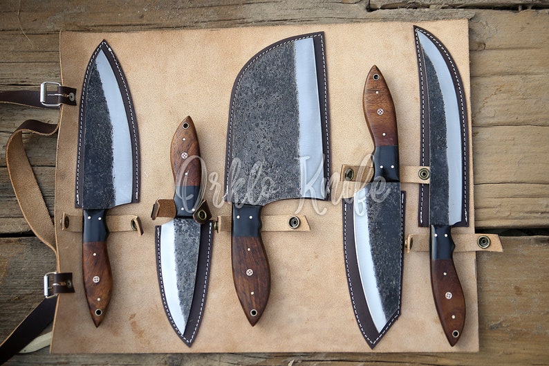 May include: Set of five kitchen knives with wooden handles and leather sheaths. The knives are arranged in a row on a wooden surface. The knives are all different sizes and shapes. The sheaths are all brown leather.