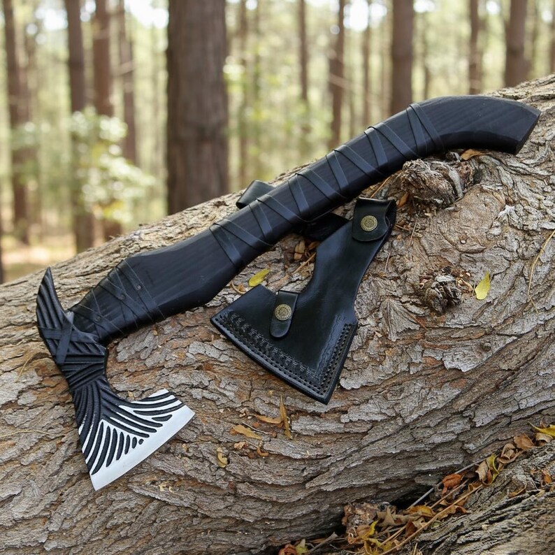 May include: A black axe with a decorative blade and a leather sheath rests on a tree trunk. The axe head features a white and black design. The handle is wrapped in black material. The sheath is black leather with a button closure.