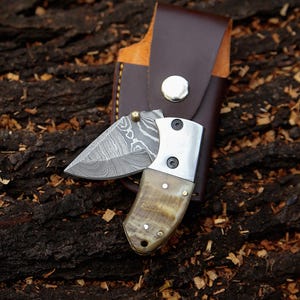 May include: A small, folding knife with a patterned steel blade and a horn handle. The knife is open, revealing the blade's intricate design. It rests on a textured wooden surface, with a brown leather sheath in the background.