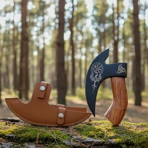 May include: A black axe with a wooden handle and a brown leather sheath. The axe head features a compass design and a decorative symbol. The sheath has two silver button closures. The axe is displayed outdoors.