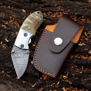 May include: A small folding knife with a patterned blade and a horn handle, next to a brown leather sheath. The knife has a silver bolster and is set against a textured wood background. The sheath has a snap closure.