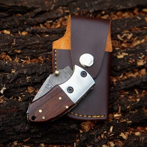 May include: A small, folding pocket knife with a dark wood handle and a silver blade, resting next to a brown leather sheath. The knife features brass rivets and a decorative pattern on the blade. The sheath has a snap closure.