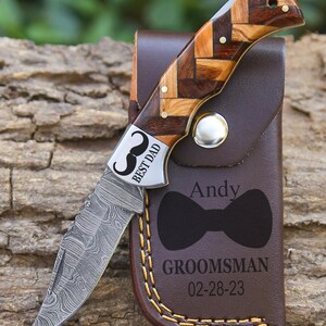 May include: A folding knife with a Damascus steel blade and a handle featuring a brown wood herringbone pattern. The bolster is engraved with a mustache and the text BEST DAD. It is paired with a brown leather sheath engraved with a bow tie, the name Andy, the word GROOMSMAN, and the date 02-28-23.